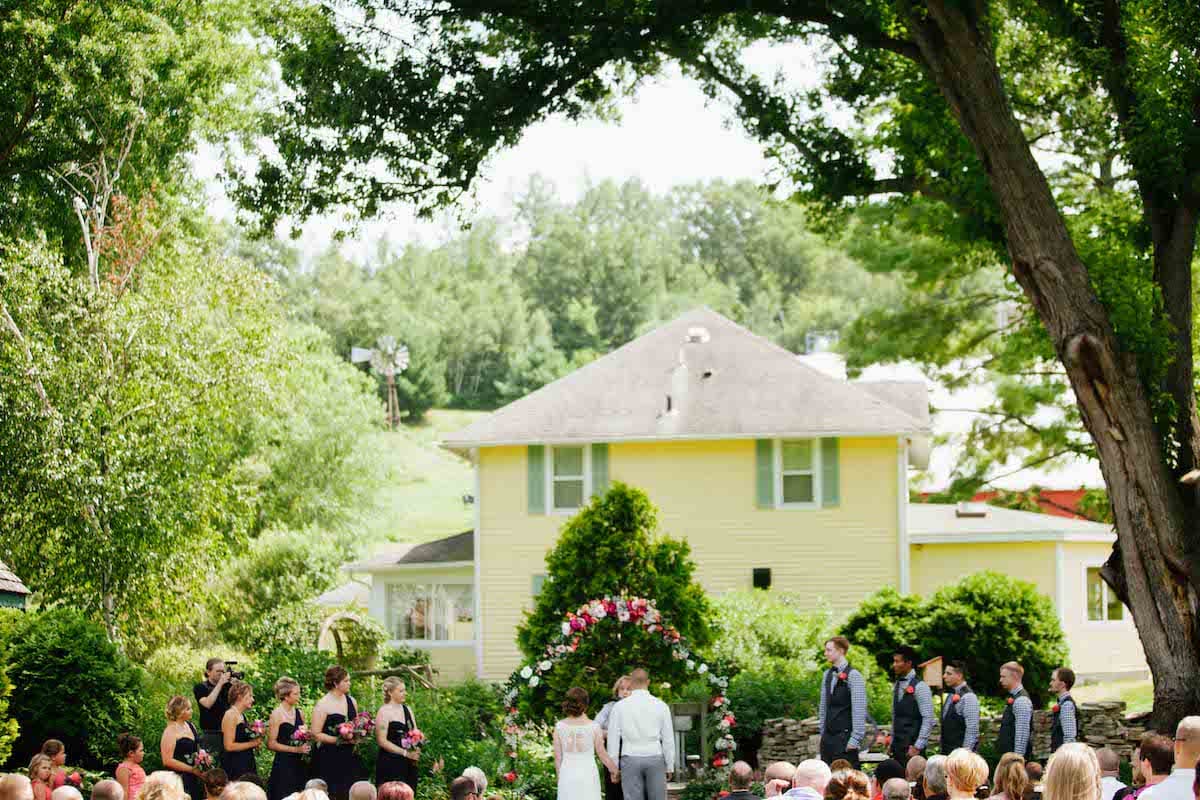 A wedding ceremony taking place outdoors with a floral arch, surrounded by guests and a yellow house in the background.