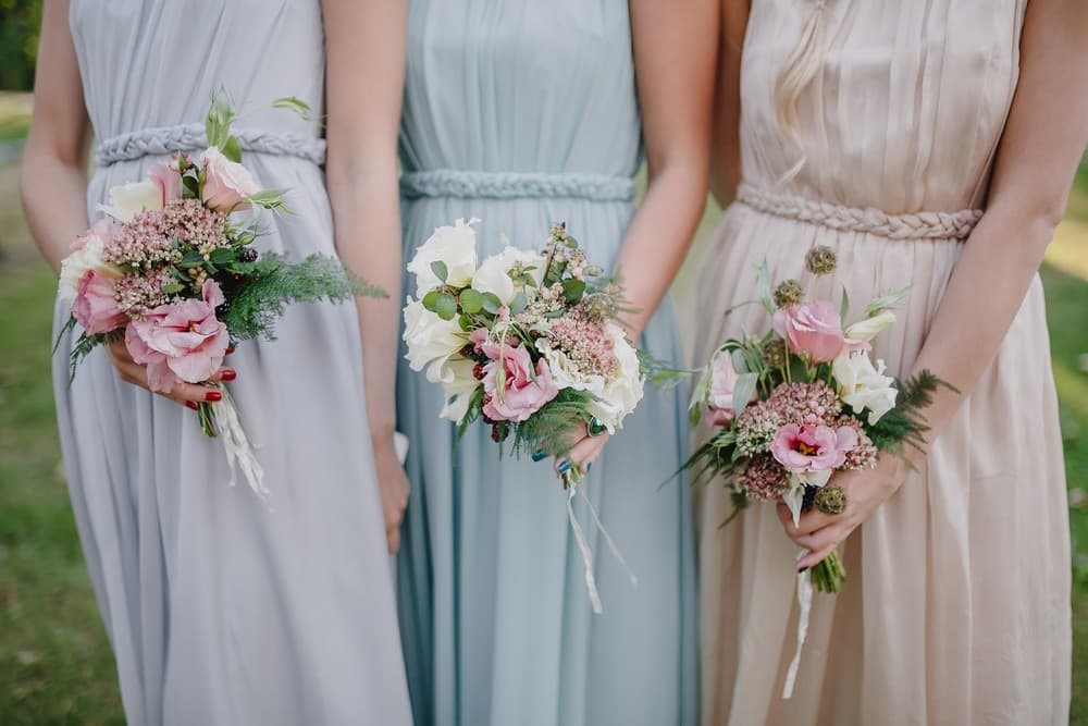 Three women in elegant dresses hold floral bouquets in a garden setting.
