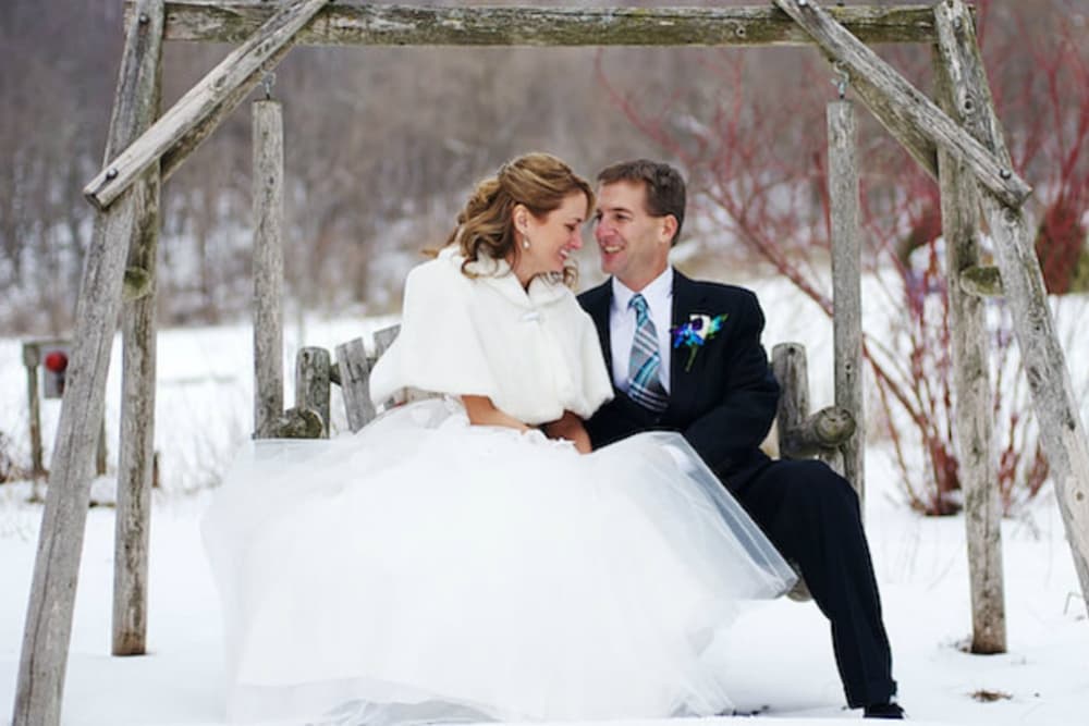A bride and groom dressed in wedding attire sitting on a wood swing outside with snow around them