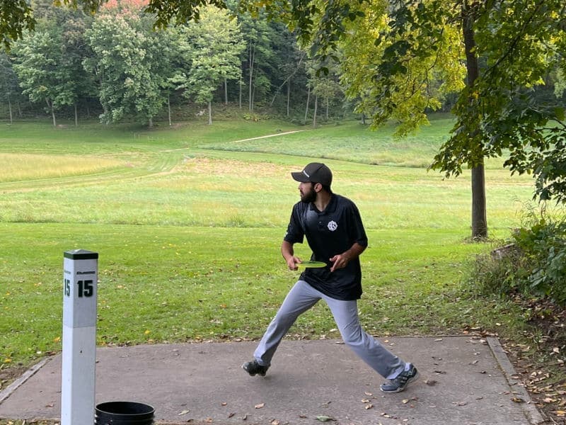 A player throws a disc from a tee pad labeled "15" on a scenic disc golf course.