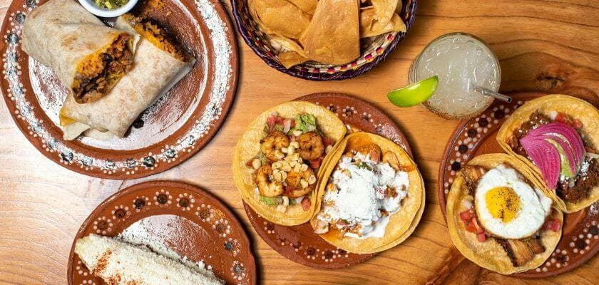 A delicious spread of tacos, a burrito, tortilla chips, and a cocktail on a wooden table.