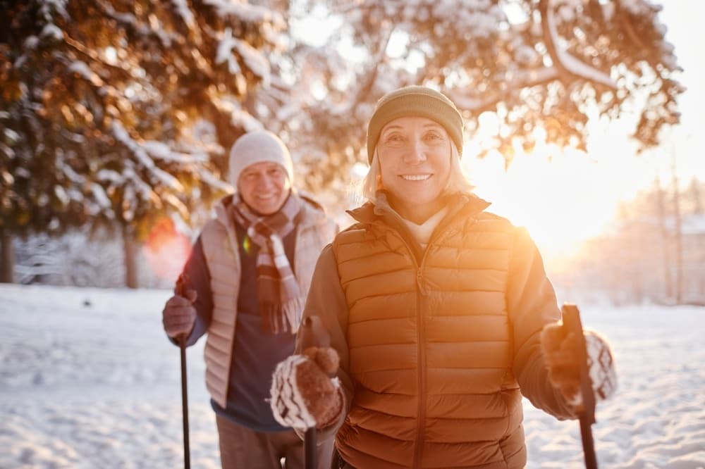 Two smiling adults dressed warmly enjoy a snowy outdoor setting during sunset.