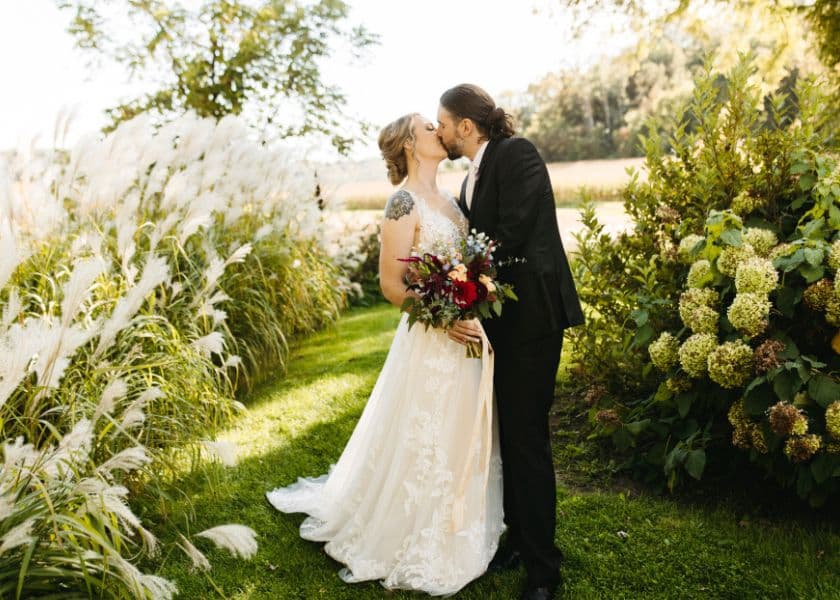 A bride and groom share a kiss surrounded by lush greenery and flowers.