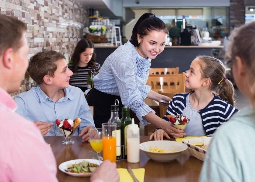 A waitress serves desserts to two children at a restaurant table surrounded by other diners. A waitress serves desserts to two children at a restaurant table surrounded by other diners.
