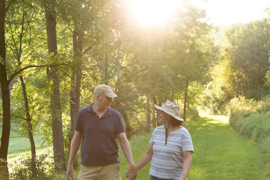 A couple walks hand-in-hand on a sunlit trail surrounded by greenery.