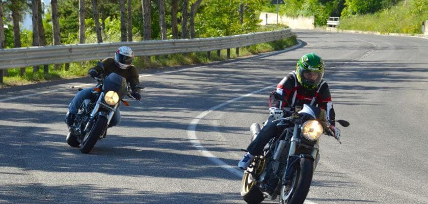 Two motorcyclists lean into a curve on a winding road surrounded by trees.