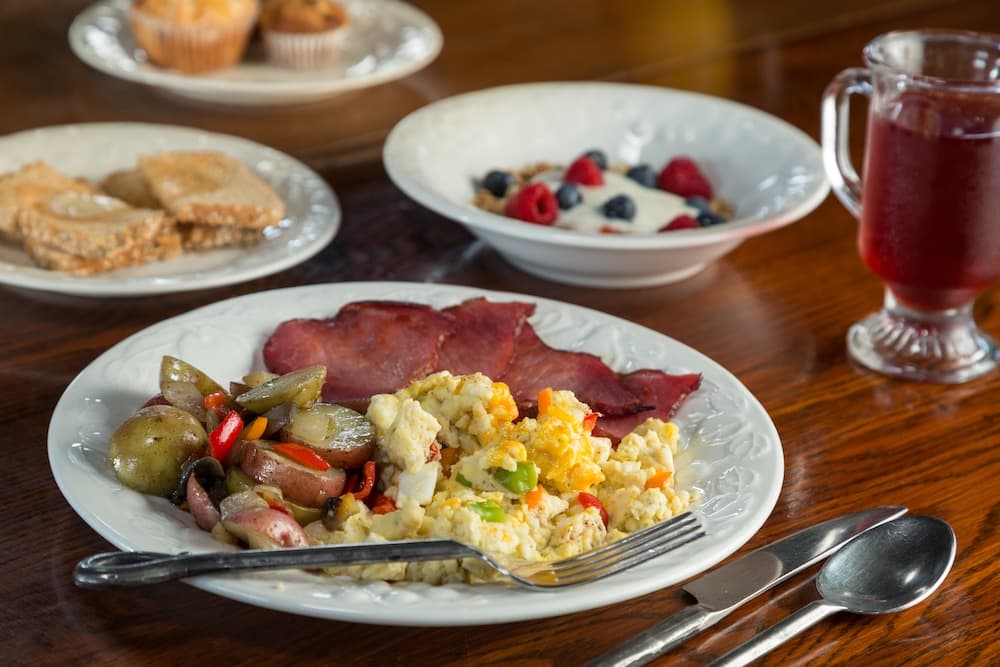 A plate of scrambled eggs, ham, and vegetables, accompanied by bowls of yogurt with berries, and a drink, with pastries in the background.