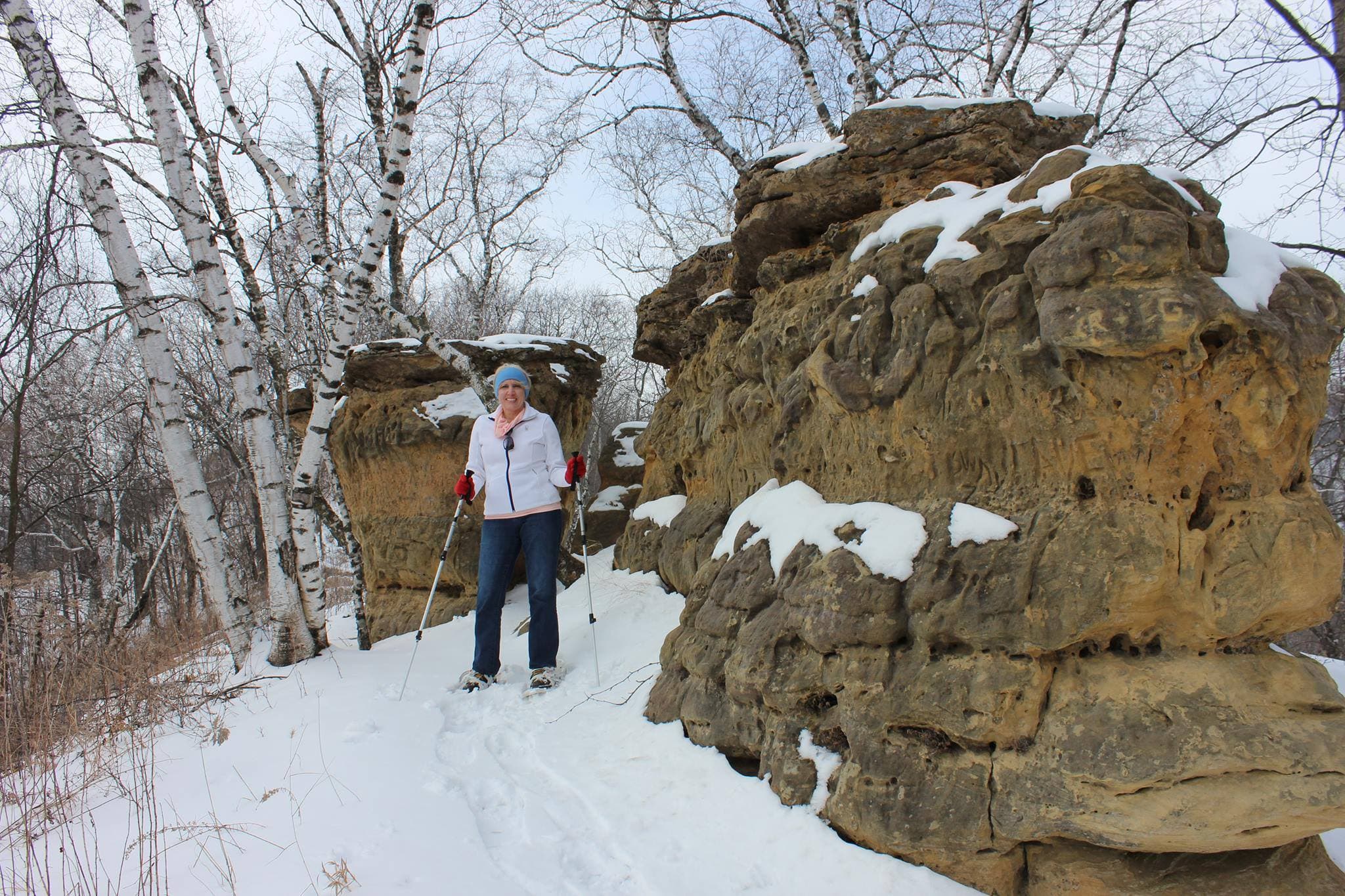 A person in winter attire stands near snow-covered rock formations in a snowy landscape with bare trees.
