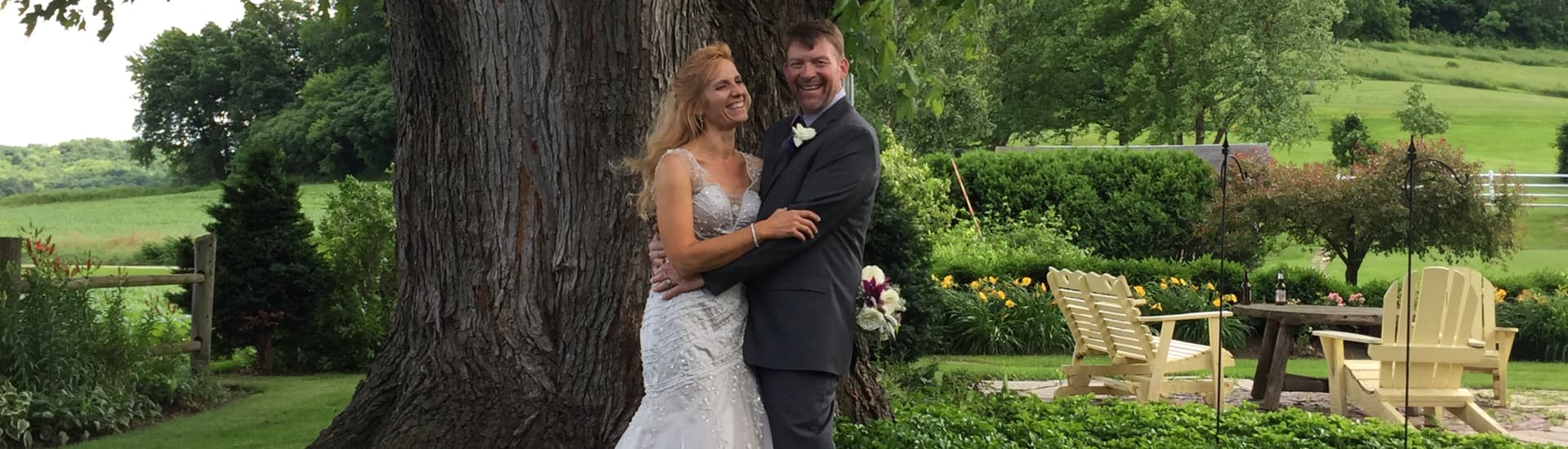A smiling couple dressed in wedding attire in front of a large oak tree, with a wood table and chairs nearby.