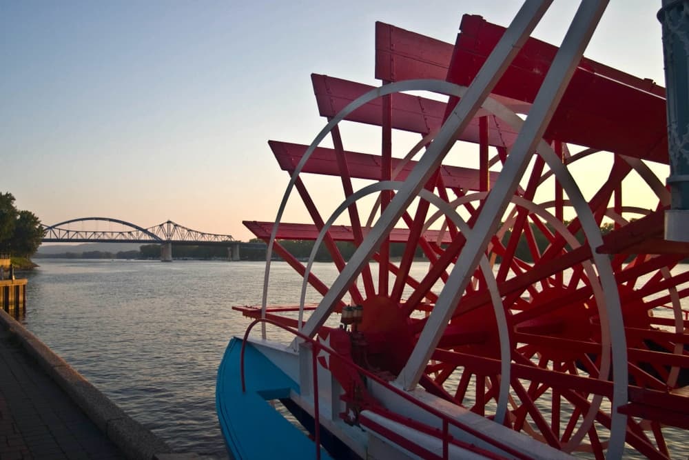 A close-up of a red paddle wheel on a riverboat with a bridge in the background at sunset.
