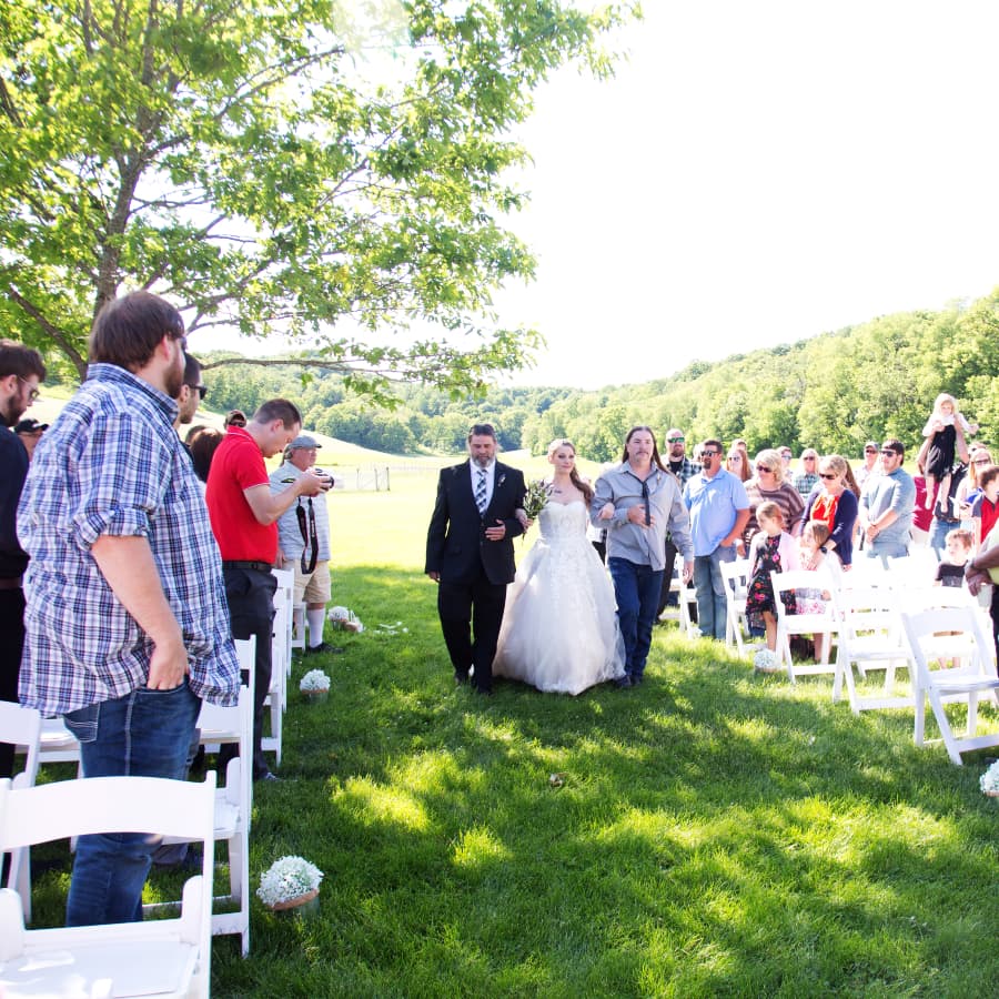 An outdoor wedding with a bride being walked down the aisle by 2 men, with guests looking on from each side.