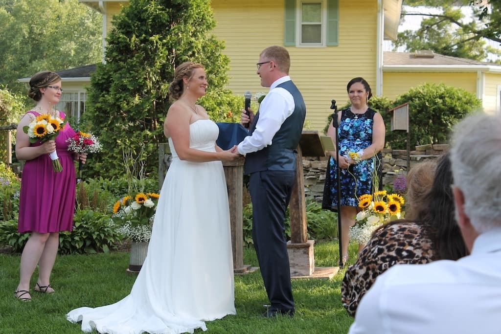 A bride and groom exchange vows during an outdoor wedding ceremony, surrounded by guests and colorful floral arrangements.