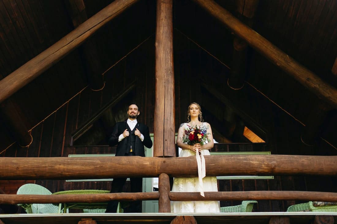 A bride and groom stand on a rustic balcony, with the groom in a suit and the bride holding a bouquet, framed by wooden beams.