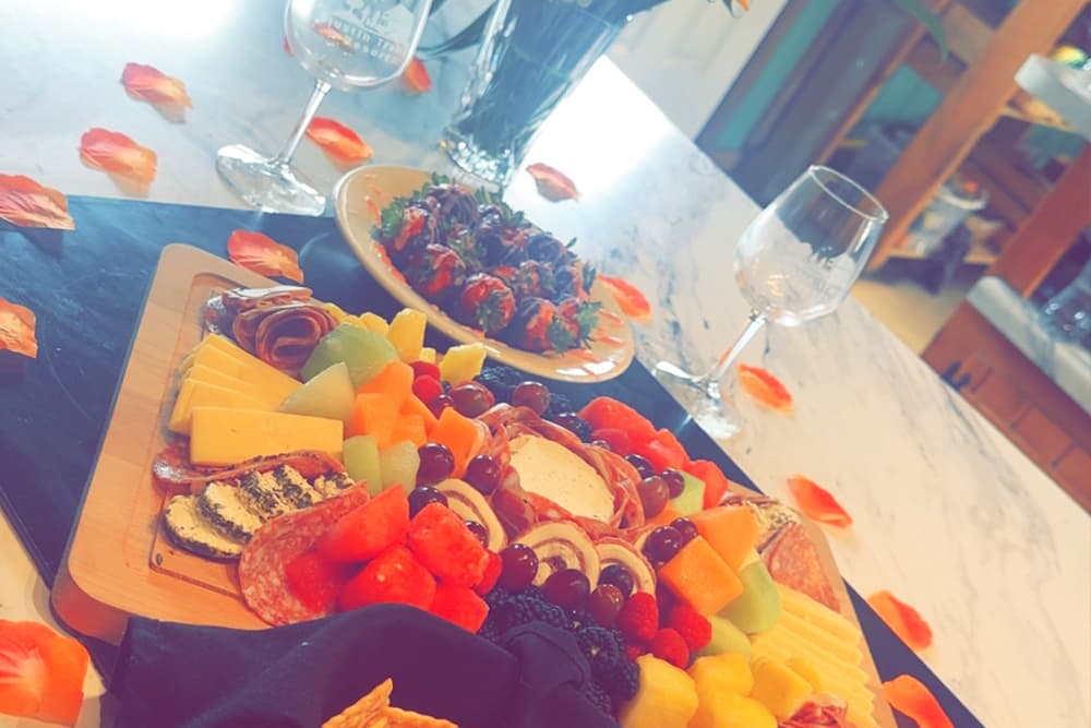 A colorful charcuterie board with assorted meats, cheeses, and fruits, alongside chocolate-covered strawberries and wine glasses, set on a white table.