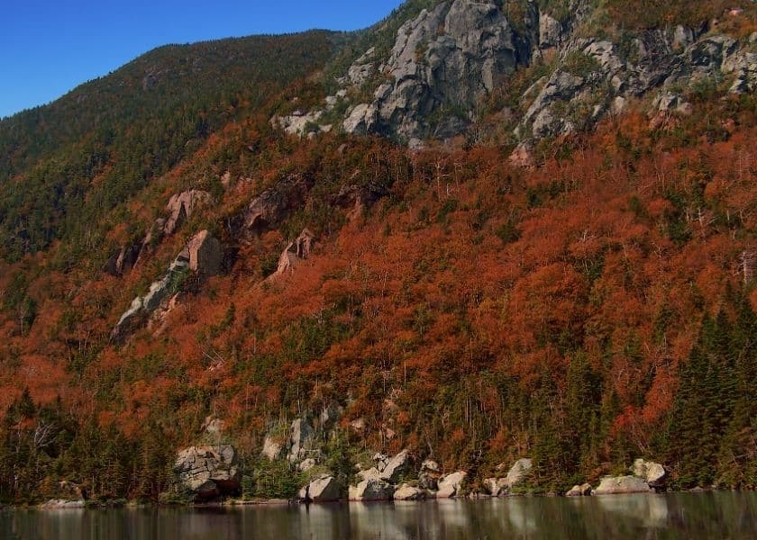 Autumn foliage reflects on a calm lake surrounded by rocky mountains.