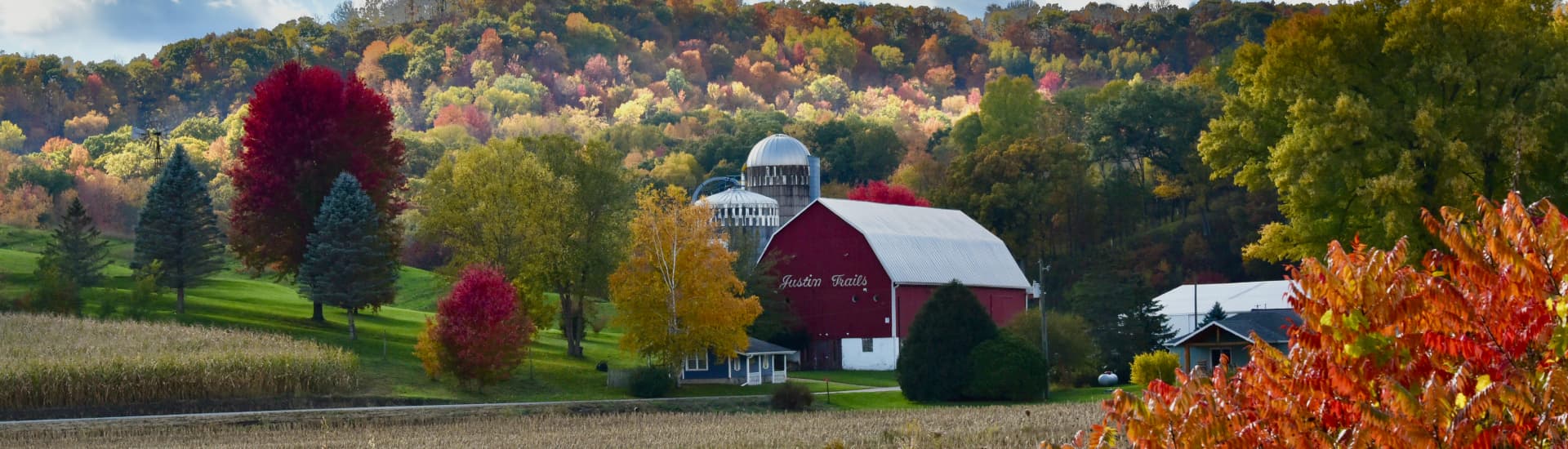 A red barn with a white roof that says Justin Trails on the front, with silos in the background, and beautiful fall colors all around colors of reds, yellows, browns and oranges.