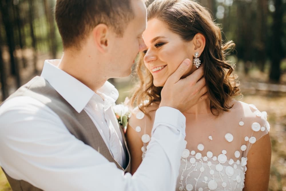 A couple shares a tender moment outdoors, with the man gently cradling the woman's face as they smile at each other.