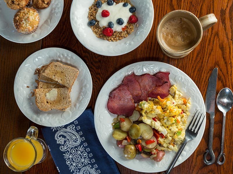 An aerial view of a breakfast table with scrambled eggs, ham slices, potatoes with peppers, a bowl of yogurt with granola and berries, buttered toast, along with cups of coffee and glasses of orange juice