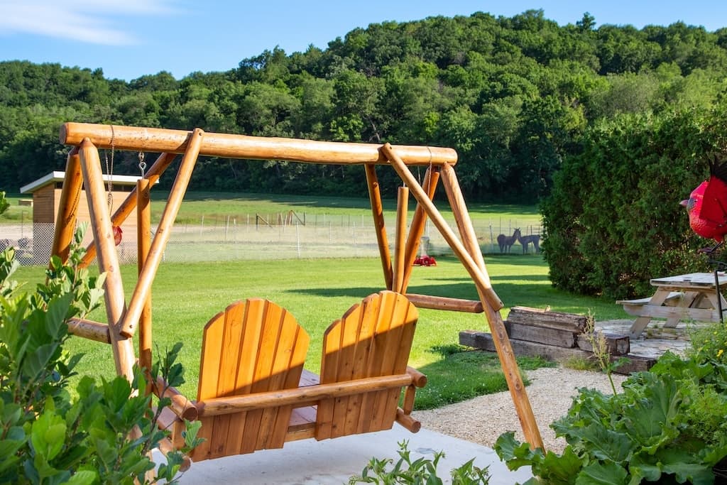 A wooden swing with two chairs overlooks a lush green landscape and distant hills.