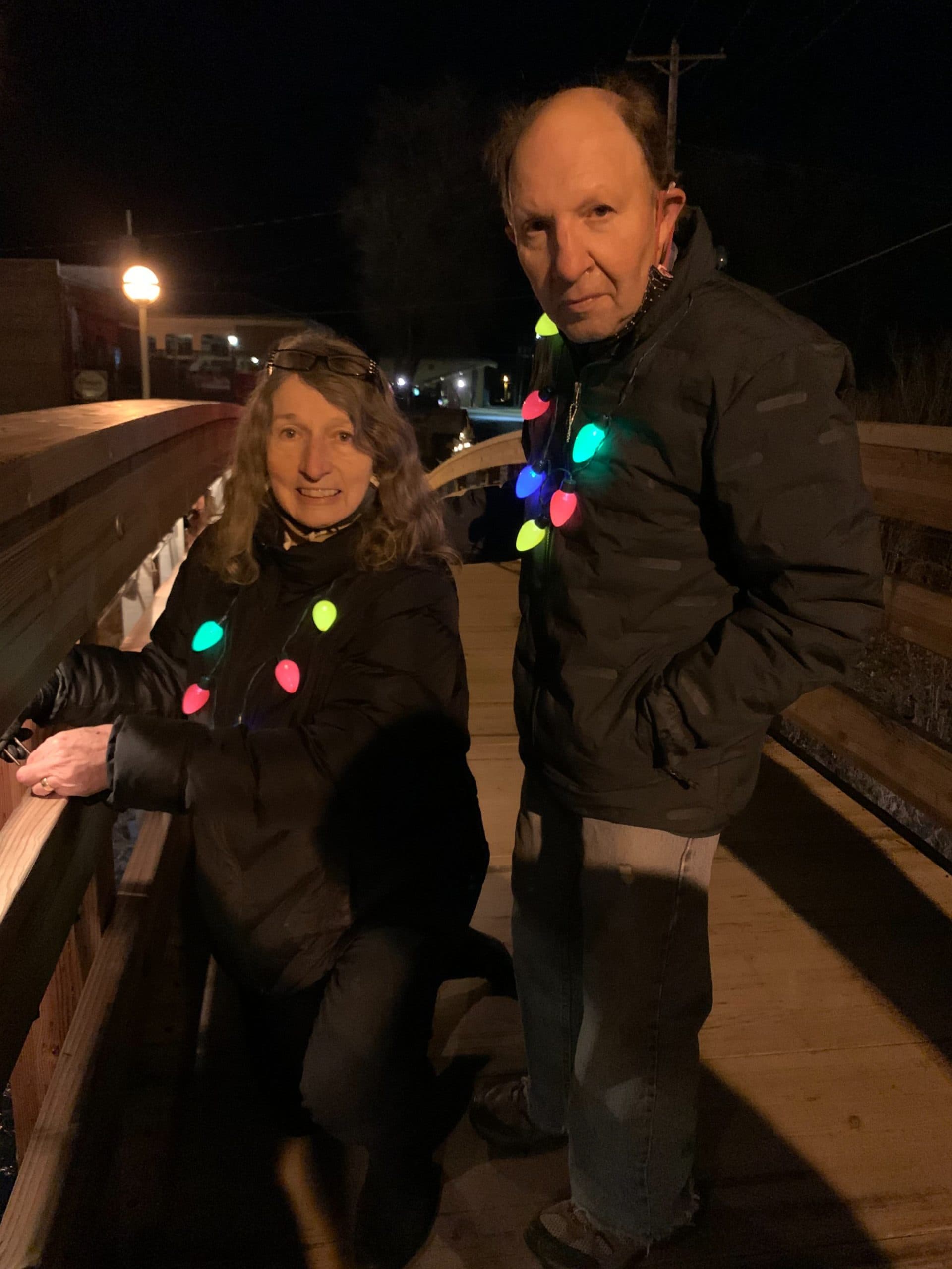 A man and woman wearing colorful holiday lights stand on a wooden bridge at night.