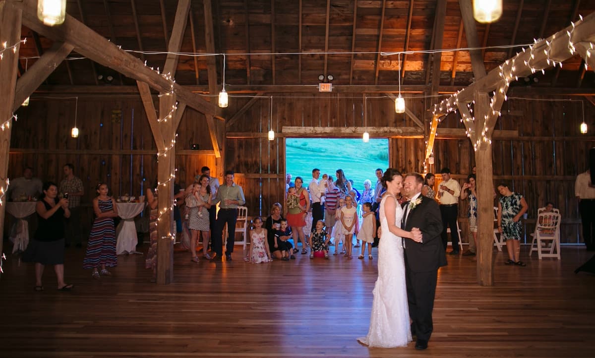 A bride and groom dance together in a warmly lit barn surrounded by guests celebrating.