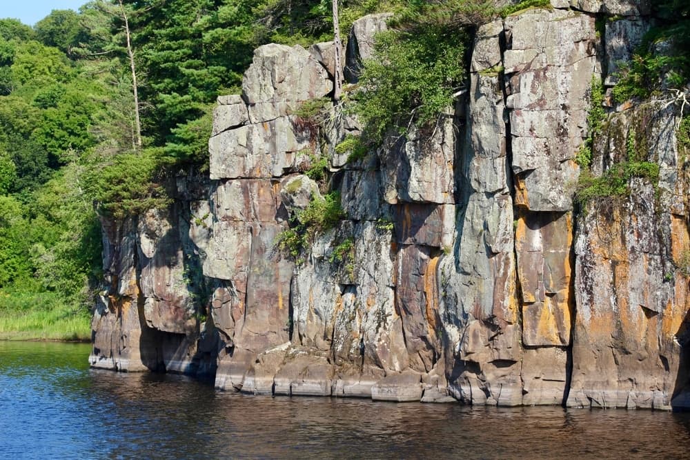 A rocky cliff rises beside a calm lake, surrounded by lush greenery.