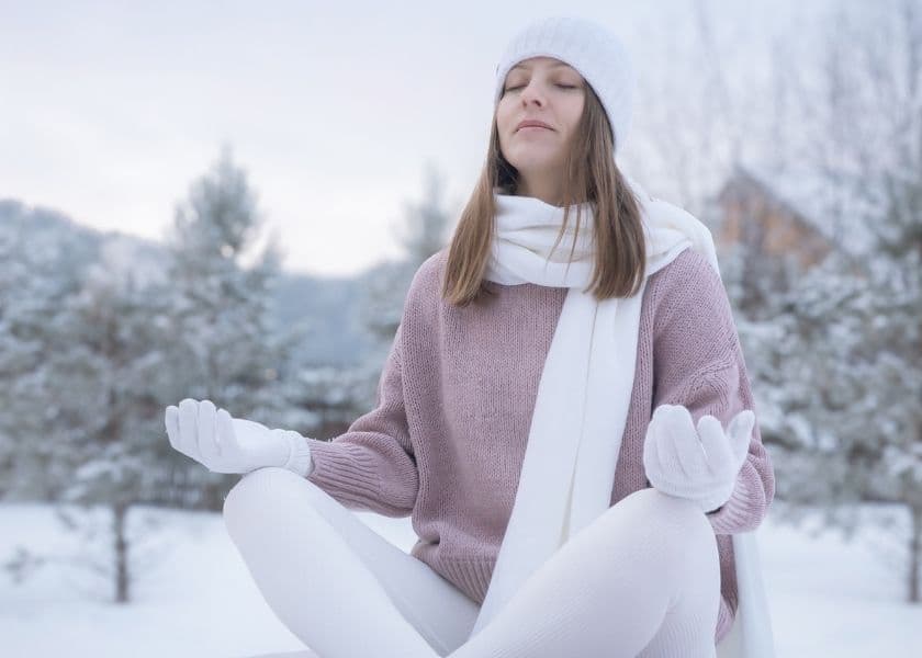 A woman in winter attire meditates peacefully in a snowy landscape.