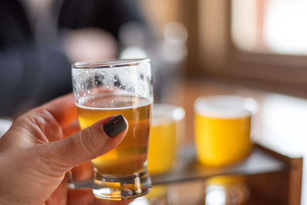 A hand holds a small glass of pale beer with blurred glasses in the background.