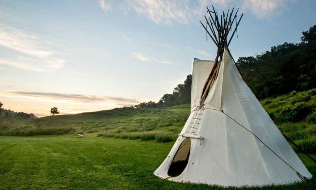 A white teepee stands in a grassy field under a cloudy sky at sunrise.