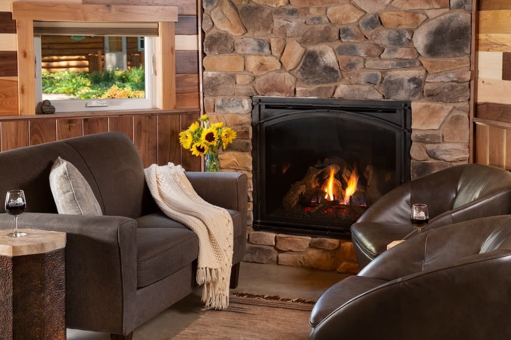Cozy living area with a stone fireplace, comfortable chairs, a side table, and sunflowers in a window.