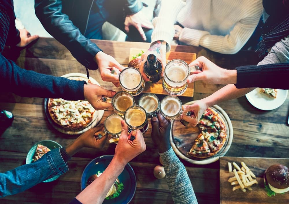A group of hands toasting with beers over a table filled with pizza and appetizers.