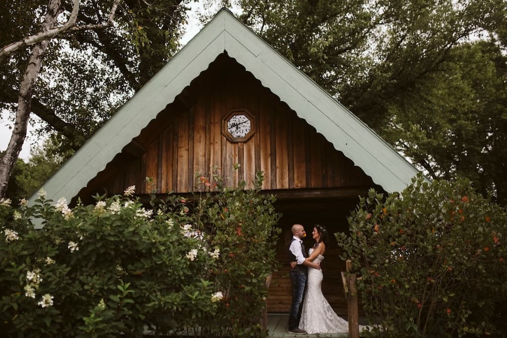 A couple embraces in front of a rustic cabin surrounded by greenery.