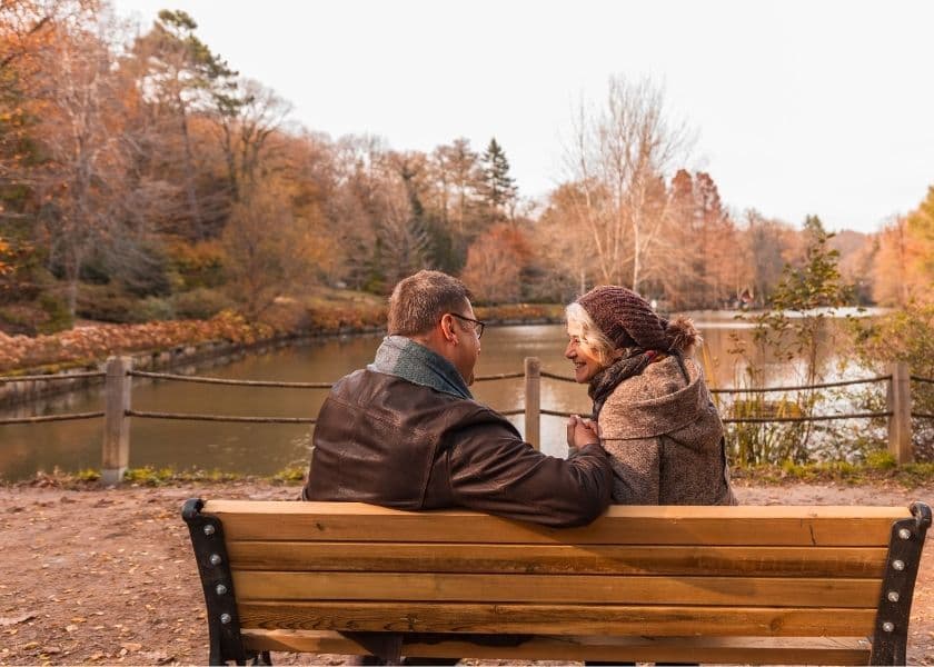 A couple sits on a bench by a serene lake, enjoying each other's company amidst autumn foliage.