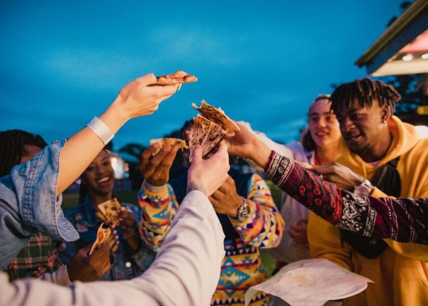 A group of friends joyfully sharing food in a lively outdoor setting during twilight. A group of friends joyfully sharing food in a lively outdoor setting during twilight.