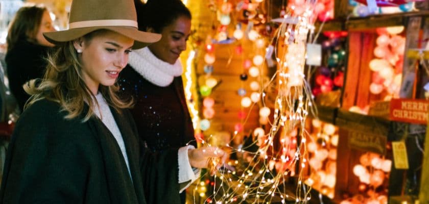 Two women admire colorful string lights at a festive market.