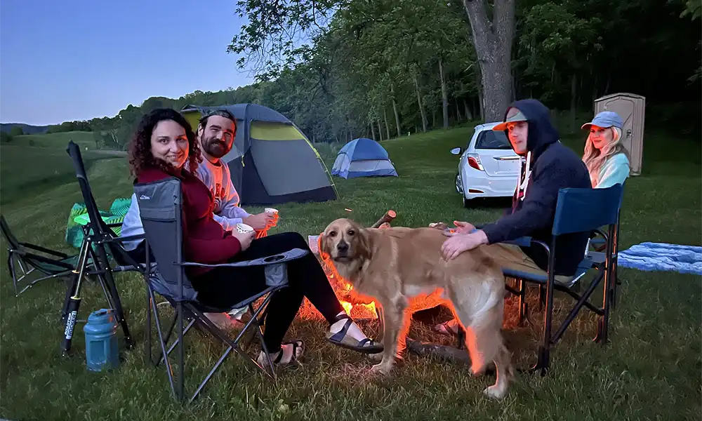 A group of four people and a golden retriever gather around a campfire as tents are set up in the background.