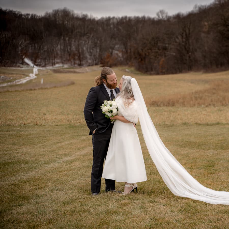 A couple dressed in wedding attire outside in a field with trees in the background.
