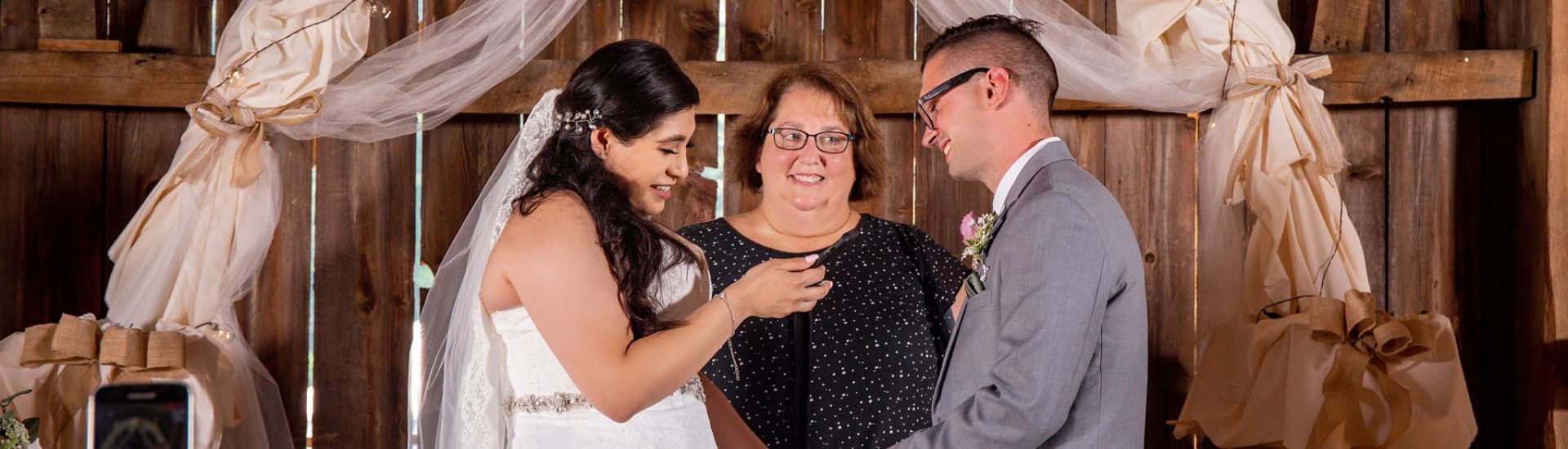 A bride, officiant, and groom share a moment during a wedding ceremony in a rustic setting.