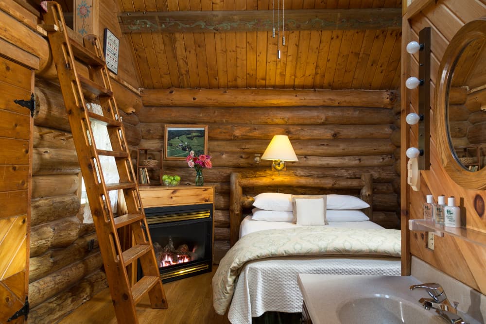 A rustic log cabin bedroom featuring a fireplace, a ladder leading to a loft, a queen-sized bed, and a vanity area with a sink and mirror.