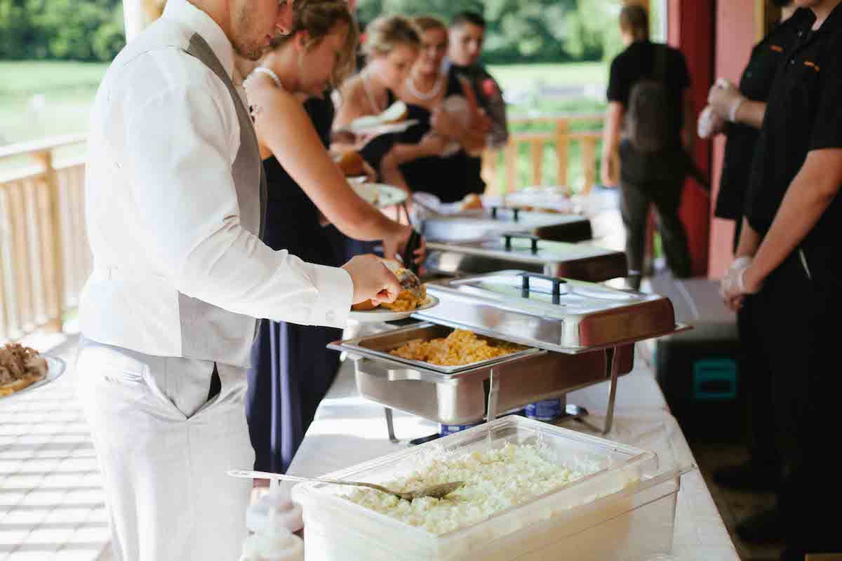 A man serves food from a buffet line while guests line up to choose their meals outdoors.