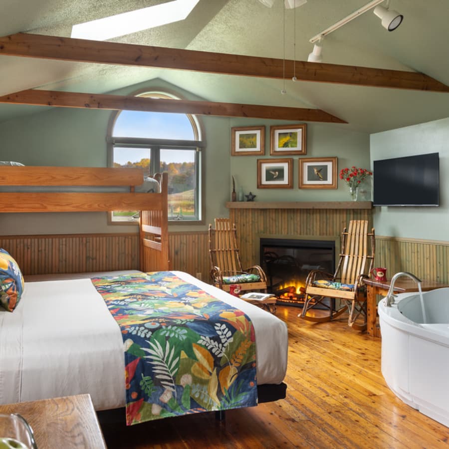 A cozy bedroom featuring a bunk bed, queen bed, fireplace, and a large tub in a cabin-like setting.