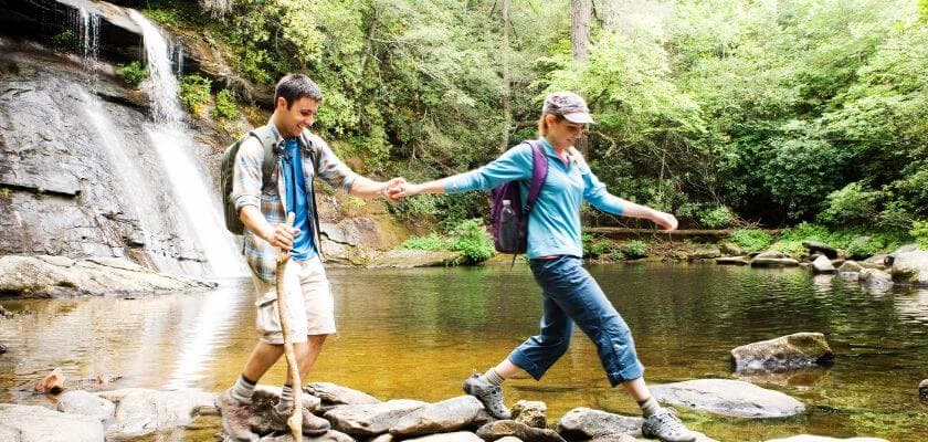 A couple walks hand-in-hand across stones in a serene, wooded area near a waterfall.