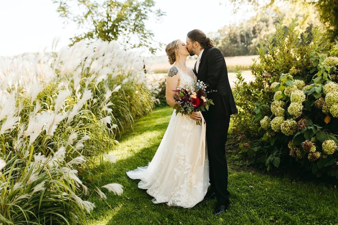 A bride and groom share a kiss in a lush garden, surrounded by greenery and soft flowers.
