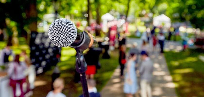 A microphone is in focus in the foreground, with a blurred crowd and event setup in a park background.