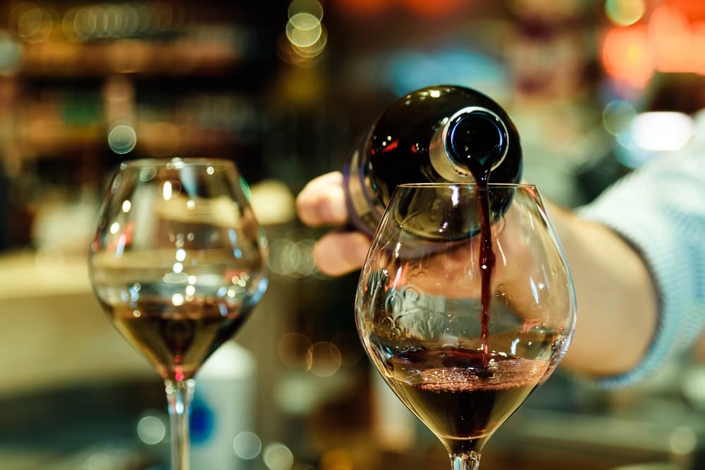 A hand pours red wine into a glass at a bar.