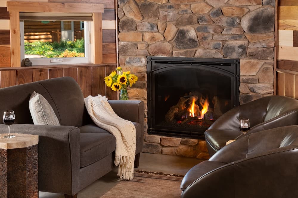 A cozy living room featuring a stone fireplace, comfortable chairs, a small table, and a vase of sunflowers.