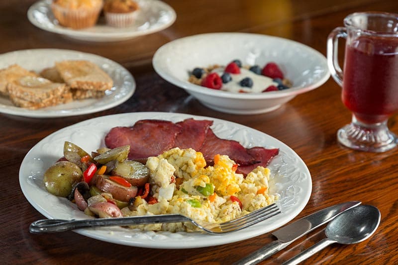 A table with a white plate filled with scrambled eggs, potatoes and peppers, ham slices, a bowl of granola with yogurt and fresh berries, sliced toast with butter, muffins, and a cup of coffee