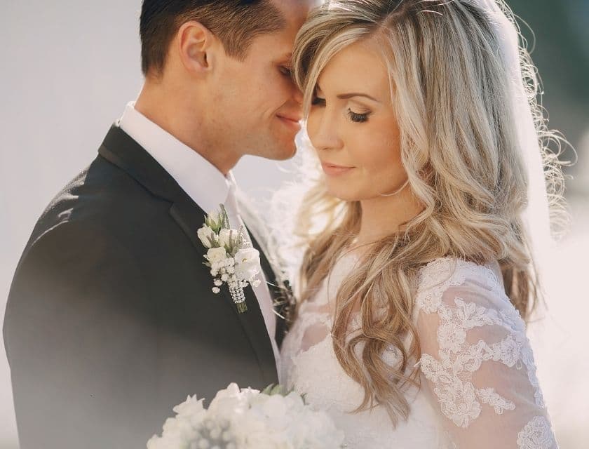 A couple tenderly embraces, with the bride in a lace gown and the groom in a dark suit, both looking blissfully content.