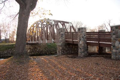 A wooden arch bridge spans a creek, surrounded by autumn leaves and trees.