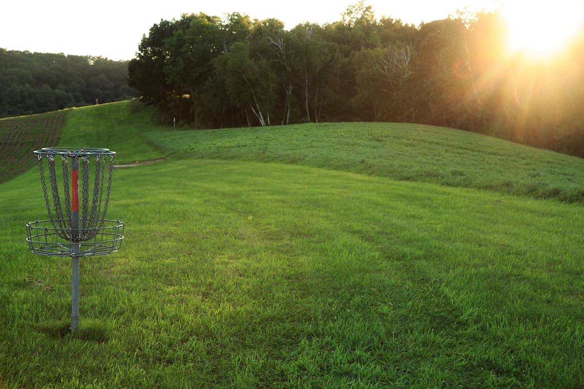 A disc golf basket stands on a lush green hillside with trees and a sunset in the background.